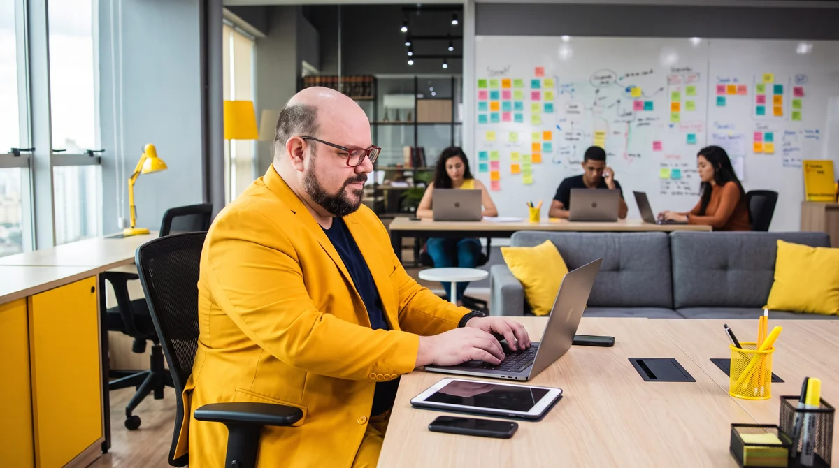 Flávio de blazer amarelo digita no notebook; sofá cinza e equipe com MacBooks diante de parede de post-its